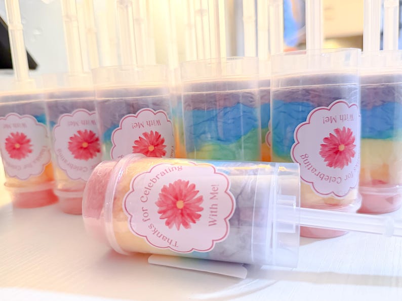 May include: Clear push-pop containers filled with colorful, layered treats. Each container has a label with a pink flower and the text "Thanks for Celebrating With Me!". The treats inside have pastel colors.