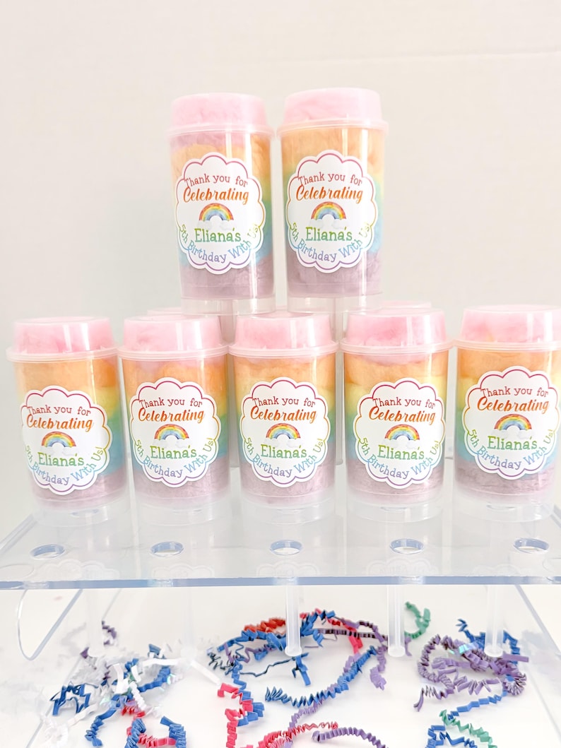 May include: A display of cotton candy party favors in clear plastic containers with pink lids. Each container has a rainbow of cotton candy and a sticker that says "Thank you for Celebrating Eliana's 5th Birthday With Us."