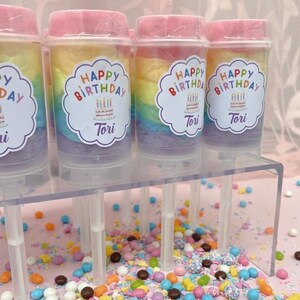 May include: Cotton candy push pops with rainbow layers and pink lids. Each pop has a label that says "Happy Birthday Tori" with a cake and candles. Colorful sprinkles are scattered on a pink surface.
