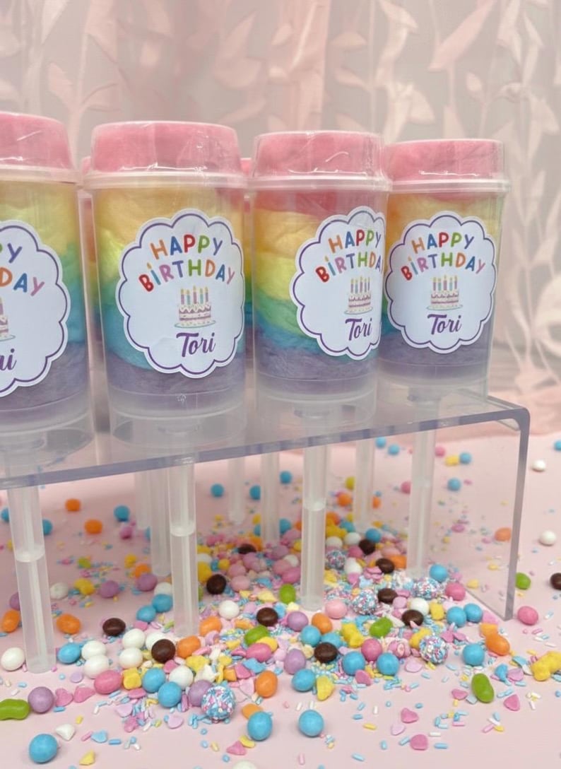 May include: Cotton candy push pops with rainbow layers and pink lids. Each pop has a label that says "Happy Birthday Tori" with a cake and candles. Colorful sprinkles are scattered on a pink surface.
