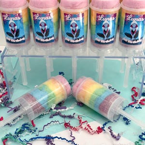 May include: Clear plastic push pop containers filled with rainbow-colored cotton candy. Each container has a pink lid and a label that reads "Luna's 8th Birthday" with a cartoon character. The containers are displayed on a clear plastic stand.