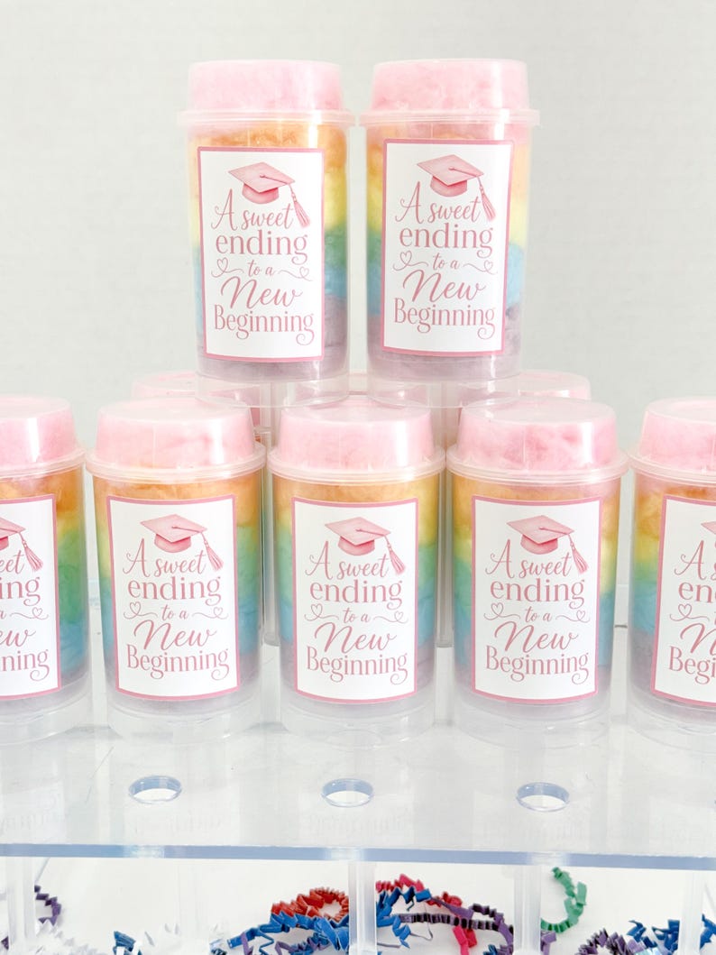 May include: Clear plastic containers filled with colorful cotton candy. Each container has a pink lid and a label with a graduation cap and the text "A sweet ending to a New Beginning."