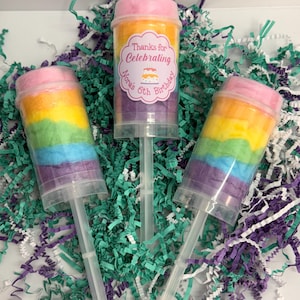 May include: Three push-pop containers filled with rainbow-colored cotton candy. One container has a label that reads "Thanks for Celebrating Nora's 6th Birthday" with a cake graphic. The containers are on a bed of teal, purple, and white shredded paper.