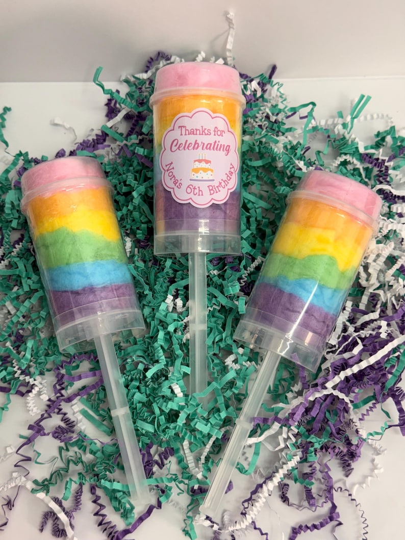 May include: Three push-pop containers filled with rainbow-colored cotton candy. One container has a label that reads "Thanks for Celebrating Nora's 6th Birthday" with a cake graphic. The containers are on a bed of teal, purple, and white shredded paper.