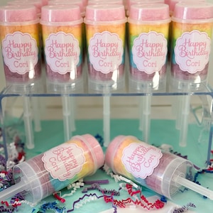 May include: A collection of push-pop containers filled with rainbow-colored cotton candy. Each container has a pink lid and a label that reads "Happy Birthday Cori". The containers are displayed on a clear acrylic stand, with two lying down.