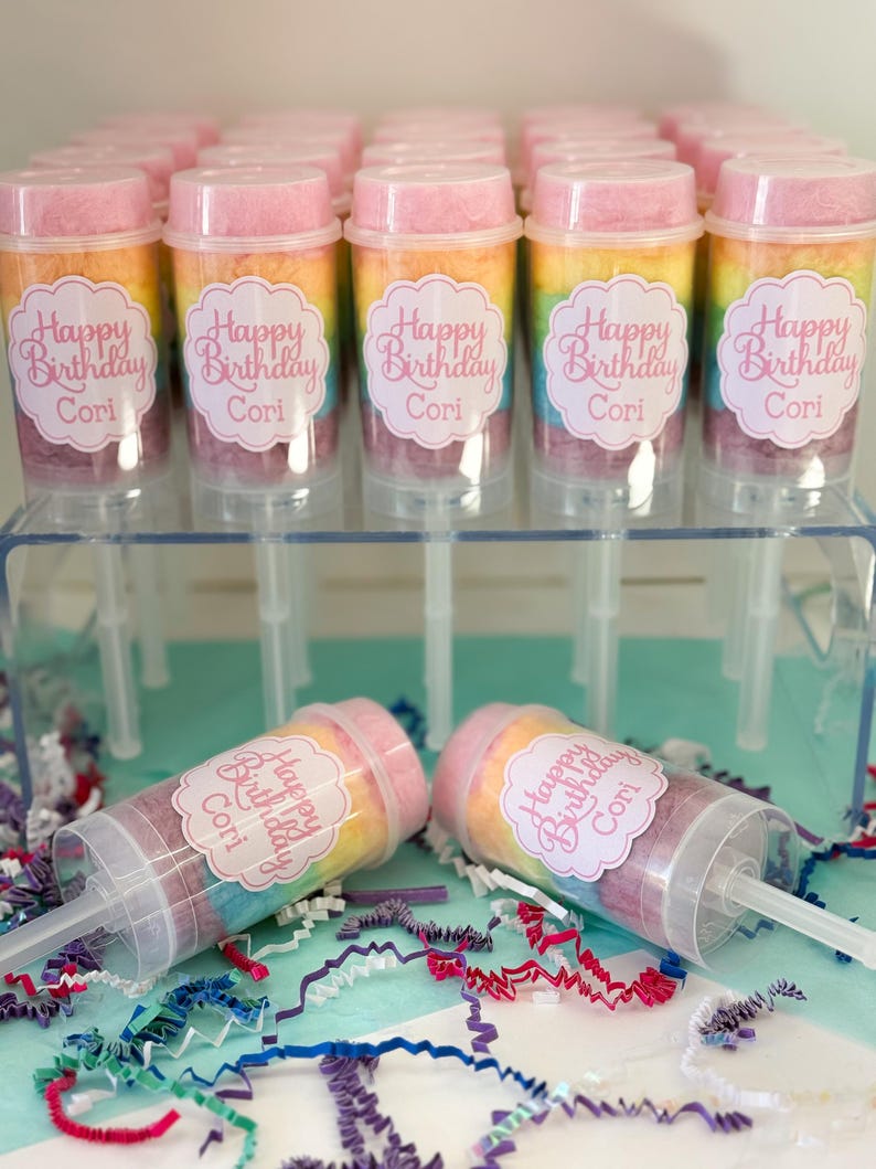 May include: A collection of push-pop containers filled with rainbow-colored cotton candy. Each container has a pink lid and a label that reads "Happy Birthday Cori". The containers are displayed on a clear acrylic stand, with two lying down.
