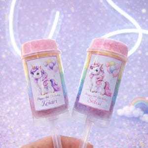 May include: Two push pop containers with pink lids and unicorn illustrations. Each container has a label with a name and birthday message. The background is a sparkly purple with a neon light.