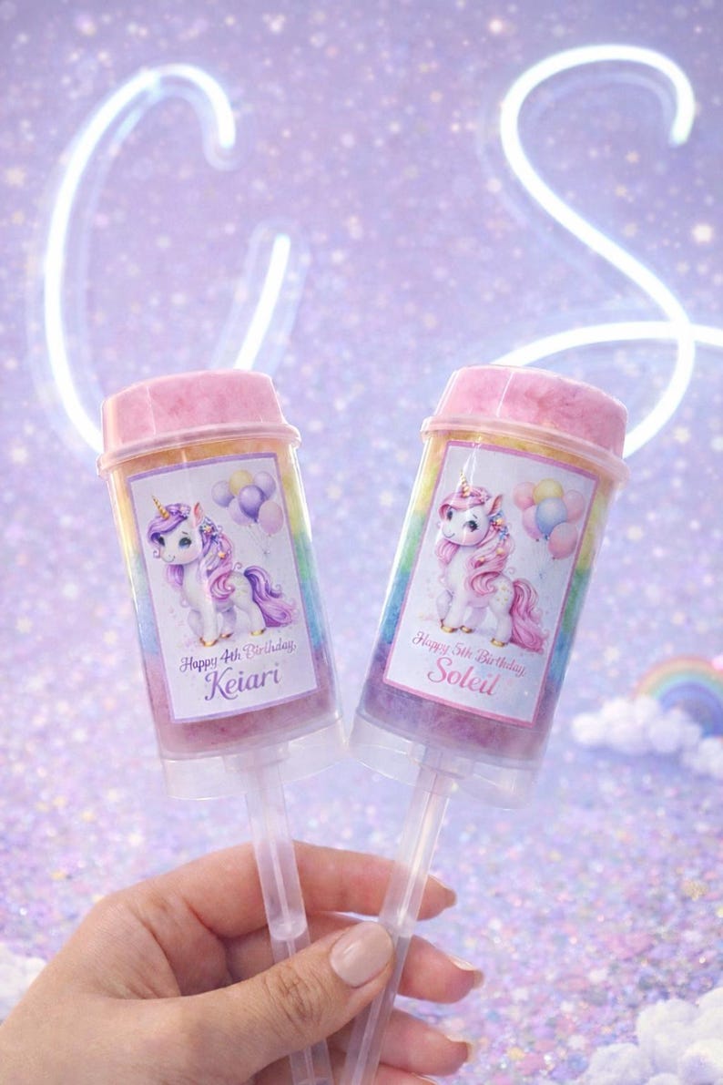 May include: Two push pop containers with pink lids and unicorn illustrations. Each container has a label with a name and birthday message. The background is a sparkly purple with a neon light.