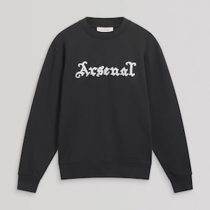 May include: Black crew neck sweatshirt with the word "Arsenal" in white, stylized lettering across the chest. The long-sleeved top has a ribbed hem and cuffs. The sweatshirt is made of a soft material.