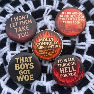 May include: Five round buttons with various phrases in white and gold lettering against dark backgrounds. The buttons have a distressed, vintage look. Phrases include "I WON'T LET THEM TAKE YOU" and "I'D WALK THROUGH HELL FOR YOU."