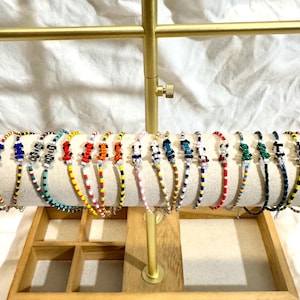 Peut inclure: Présentation de bracelets de perles multicolores, notamment bleus, rouges, jaunes et noirs. Les bracelets sont disposés sur un présentoir doré avec un support cylindrique crème. Les bracelets présentent un design délicat sur fond blanc.