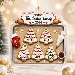 May include: A clear acrylic Christmas ornament shaped like a baking sheet with a wooden rolling pin topper. It features the text The Carter Family 2026, five tree-shaped cookies labeled Noah, Mia, Oliver, Ava, and Lucy, a red mitten, and a piping bag.