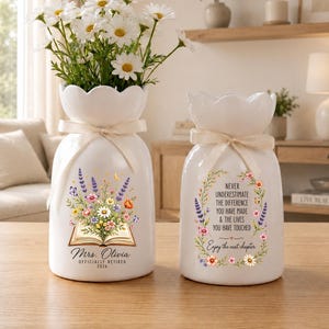 May include: Two white ceramic vases with scalloped rims and cream-colored ribbons. The left vase features a floral arrangement emerging from an open book with the text Mrs. Olivia Officially Retired 2026. The right vase displays a floral wreath with the text Never Underestimate The Difference You Have Made & The Lives You Have Touched Enjoy the next chapter.