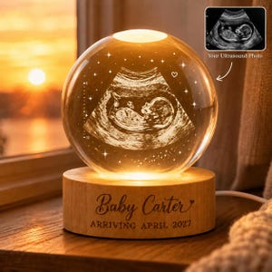 May include: A clear glass sphere featuring an internal 3D laser-etched ultrasound image of a fetus, illuminated by a light-up wooden base. The base is engraved with the text Baby Carter ARRIVING APRIL 2027 in a dark brown script and sans-serif font.