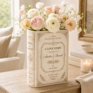 May include: A cream-colored book-shaped vase featuring the text A LOVE STORY, WRITTEN BY AMELIA & BENNETT, OUR GREATEST CHAPTER, EST. 08.16.2026, and EVERY PAGE LEADS US HOME. The vase is filled with a bouquet of white, cream, and soft pink flowers.