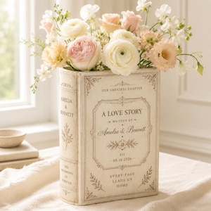 May include: A cream-colored rectangular book-shaped vase featuring the text A LOVE STORY, WRITTEN BY Amelia & Bennett, OUR GREATEST CHAPTER, EST. 08.16.2026, and EVERY PAGE LEADS US HOME, filled with a bouquet of white, cream, and soft pink flowers.