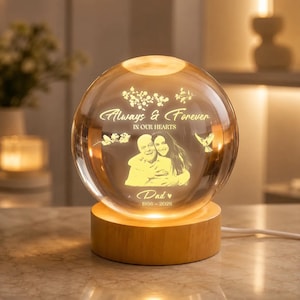 May include: A glowing crystal ball with a wooden base. The sphere features an etched image of a couple and the words "Always & Forever In Our Hearts" and "Dad 1956-2026". The base is made of light-colored wood.