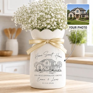 May include: A white ceramic vase with a scalloped rim, tied with a gold ribbon. It features a black line drawing of a house, the text "Home Sweet Home", "248 WILLOWBROOK DRIVE", "Emma & Lucas", and "11.14.2026". The vase is filled with white baby's breath.
