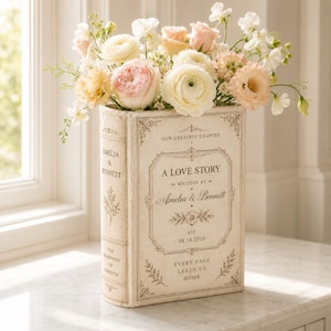 May include: A rectangular cream-colored book-shaped vase filled with a bouquet of pale pink, peach, and white flowers. The front features text reading A LOVE STORY, WRITTEN BY Amelia & Bennett, EST. 08.16.2026, and EVERY PAGE LEADS US HOME.