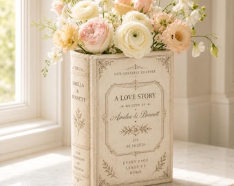 May include: A rectangular cream-colored book-shaped vase filled with a bouquet of pale pink, peach, and white flowers. The front features text reading A LOVE STORY, WRITTEN BY Amelia & Bennett, EST. 08.16.2026, and EVERY PAGE LEADS US HOME.