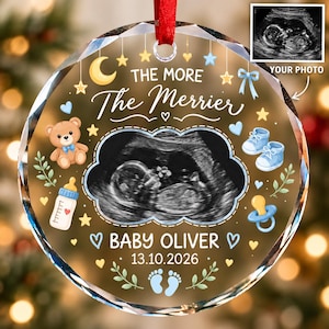 May include: A round, faceted glass ornament featuring a central ultrasound image, the text THE MORE The Merrier, BABY OLIVER, and 13.10.2026. Graphics include a teddy bear, baby shoes, a bottle, a pacifier, stars, hearts, and footprints in blue and yellow.