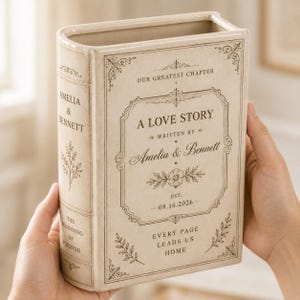 May include: A cream-colored ceramic book-shaped vase featuring dark brown text and floral illustrations. The front reads A LOVE STORY, WRITTEN BY Amelia & Bennett, OUR GREATEST CHAPTER, EST. 08.16.2026, and EVERY PAGE LEADS US HOME. The spine reads AMELIA & BENNETT.