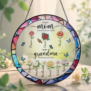 May include: A round, stained-glass window art piece with a black chain for hanging. The design features the words "First Mom Growing Since 1995" and "Now Grandma Growing Since 2015." It includes floral and butterfly motifs, with names like Mason, Daniel, and Nicole.
