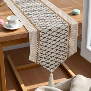 Peut inclure: Chemin de table crème avec un motif de losanges noirs. Le chemin de table a une texture tissée avec une bordure beige et un pompon blanc. Une tasse et une soucoupe blanches sont sur la table en bois.