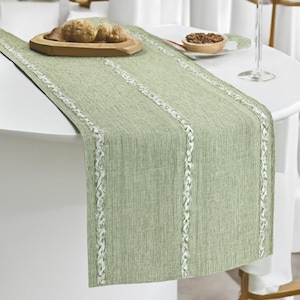 May include: A sage green table runner with three vertical white braided trim details. The runner is draped over a white table, with a wooden serving dish and small bowl visible on top. The runner adds a touch of elegance to any dining setting.