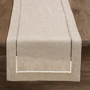 Puede incluir: Camino de mesa de lino beige con un borde rectangular calado. El camino de mesa está colocado sobre una mesa de madera. El borde calado está formado por pequeños agujeros espaciados uniformemente. La tela de lino tiene un aspecto texturizado.