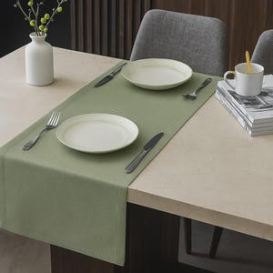 Puede incluir: Una mesa de comedor con un camino de mesa verde salvia, dos platos blancos con un fino borde dorado, cubiertos y una taza blanca con un borde dorado. También hay un jarrón blanco con tallos verdes.