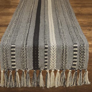 May include: A woven table runner with a striped pattern in shades of gray, black, and cream. The design features vertical stripes of varying widths and textures, with a central cream-colored stripe. The runner is finished with a fringe of tassels.