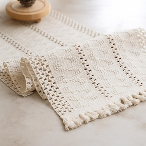 Puede incluir: Camino de mesa tejido de color crema con un patrón texturizado y detalles de flecos. El camino de mesa está doblado, mostrando el intrincado diseño y los flecos decorativos al final. Un objeto de madera está en el fondo.