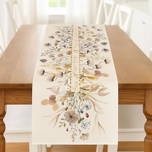 May include: Cream-colored table runner with a floral design. The runner features a central vertical band with a repeating pattern and is adorned with watercolor-style flowers in muted tones of pink, brown, and green. The runner is placed on a wooden table.