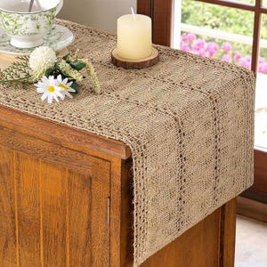 May include: A tan burlap table runner with a decorative open weave pattern. A white candle sits on a wooden coaster. A teacup and saucer, along with white flowers, are on the wooden cabinet. The runner is approximately 60 inches long.