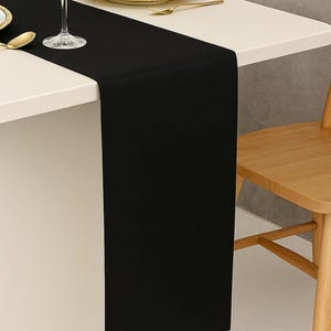Puede incluir: Un camino de mesa negro sobre una mesa blanca, con un plato con borde dorado, una copa de vino y una cuchara dorada. Una silla de madera es visible en el fondo. El camino de mesa añade elegancia.