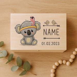 Op de afbeelding: Lichte houten kist met een cartoon koala-illustratie met een hoofdband en veer. De kist heeft het woord "NAME" en de datum "01.02.2023" erop gedrukt, samen met twee kleine vogelillustraties.