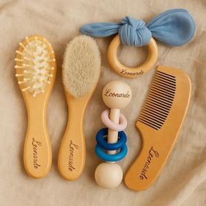 May include: A wooden baby grooming set featuring two brushes, a comb, a rattle, and a teething ring. The set includes a brush with soft bristles, a brush with wooden bristles, a comb, a rattle with colorful rings, and a blue fabric teething ring. The name "Leonardo" is engraved on each item.