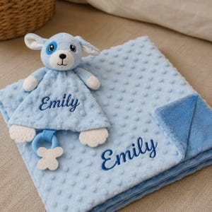 Op de afbeelding: Een lichtblauw baby-knuffeldoekje en een bijpassende pluche hond, beide voorzien van de naam Emily geborduurd in donkerblauw schrift. Het dekentje heeft een textuur met nopjes en een gladde blauwe onderkant, inclusief een witte bijtring.