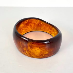 Peut inclure: Un bracelet épais de forme ovale, dans les tons marron et ambre. La matière translucide révèle un motif marbré, créant un effet écaille de tortue. Un accessoire audacieux, adapté à différents styles.