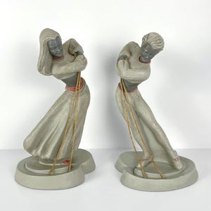 Peut inclure: Deux figurines en céramique grises représentant des femmes en robes fluides, chacune avec une chaîne dorée. Les figurines ont les cheveux foncés et sont posées dans une posture gracieuse et penchée. Les socles sont d'un gris plus clair.