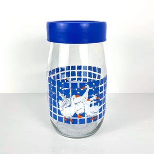May include: A clear glass jar with a blue lid. The jar has a blue and white grid pattern with white geese illustrations. The jar is cylindrical with a wide opening.