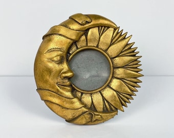 May include: A circular gold-toned picture frame featuring a sculpted crescent moon with a human face on the left side and a detailed sunflower with radiating petals on the right side, surrounding a central circular opening for a photograph.