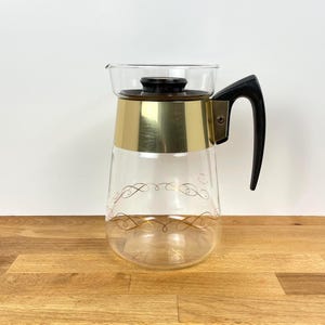 May include: A vintage glass coffee jug with a gold-coloured metal band and a black handle. The jug has a clear glass body with a decorative gold swirl design and a clear glass lid.
