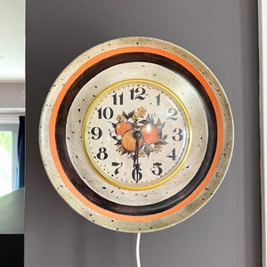 May include: A round wall clock with a speckled cream frame, a bright orange inner ring, and a dark brown border. The clock face features black numerals, a central illustration of three oranges with green leaves, and a white power cord extending downward.