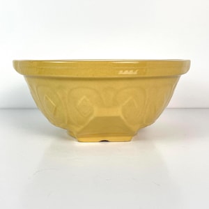 May include: A yellow ceramic mixing bowl with a geometric pattern around the sides. The bowl has a wide rim and a square base. The bowl is a solid yellow colour and is likely used for baking or cooking.