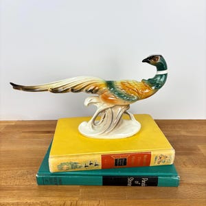 May include: A ceramic pheasant figurine featuring a glossy finish with a dark green head, a white neck ring, and a body transitioning from orange to cream. The bird has a long, sweeping tail and is perched on a cream-coloured base.
