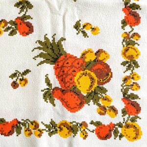 May include: A white fabric textile featuring a cross-stitch pattern of various fruits including pineapples, pears, and cherries in shades of orange, yellow, and olive green. The design includes a decorative border of fruit clusters and leaves.