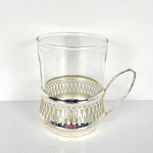 May include: A clear glass cup is held in a decorative silver-coloured metal holder with a handle. The holder features a detailed openwork design around the cup's base. The cup and holder are on a white surface.