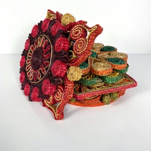 May include: A decorative hat crafted from woven straw. The hat showcases a circular design with red, burgundy, and gold accents. It features spiral and floral patterns in shades of green, orange, and yellow.